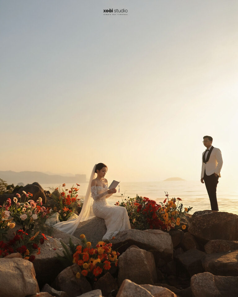 Cô dâu ngồi giữa hoa trên bãi đá, chú rể đứng phía sau bên biển Hòn Đỏ.