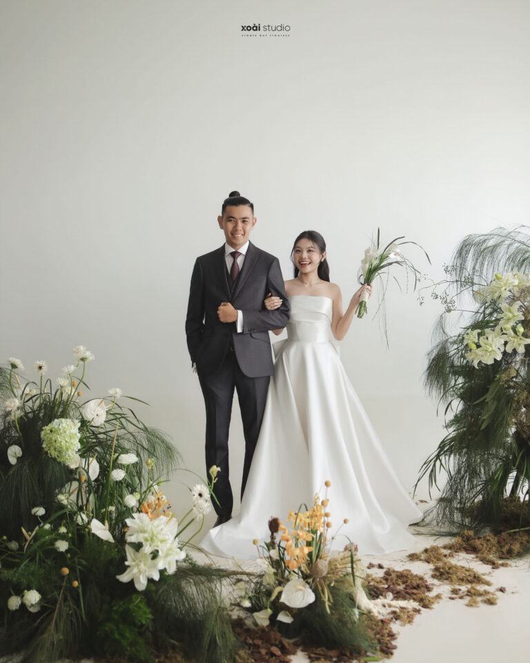 Anh và Thịnh đứng cạnh nhau giữa phông nền sáng và cụm hoa.